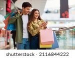 Buyers Couple Shopping Using Cellphone Holding Colorful Shopper Bags Standing In Mall. Happy Customers Using Application Purchasing Clothes Online Via Smartphone. Ecommerce And Shopaholism