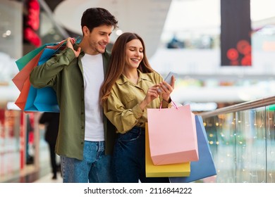 Buyers Couple Shopping Using Cellphone Holding Colorful Shopper Bags Standing In Mall. Happy Customers Using Application Purchasing Clothes Online Via Smartphone. Ecommerce And Shopaholism - Shutterstock ID 2116844222