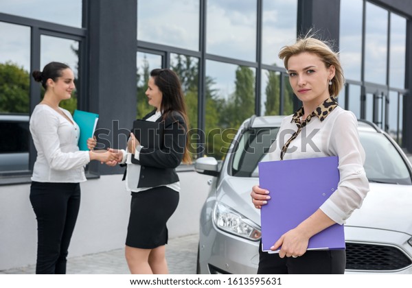 Buyers looking at car buy contract\
near new car. They have big folders in hands with\
documents