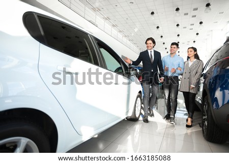 Buying Auto Concept. Successful Car Seller Helping Young Couple Choose New Automobile In Dealership Showroom