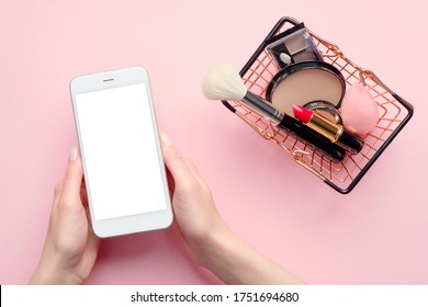 Buying Beauty Products Online Concept. Female Hand Holding Mobile Phone With Blank Screen Mockup And Shopping Cart With Cosmetics On Pink Background Top View.