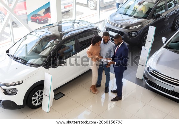 Buying Car. Auto Showroom Manager Consulting Young\
Black Spouses, Happy African American Couple Purchasing New Auto In\
Modern Dealership Center, Listening Vehicle Characteristics, Top\
View Shot