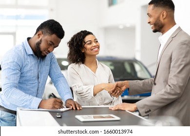 Buying Car. Black Couple Shaking Hands With Salesman And Signing Papers In Cars Selling Center. Selective Focus