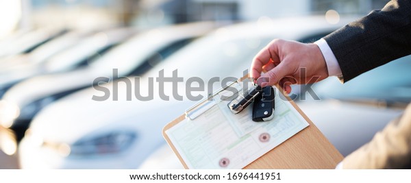 \
Buying a car, seller holds registration\
papers and keys in front of a row of new\
cars