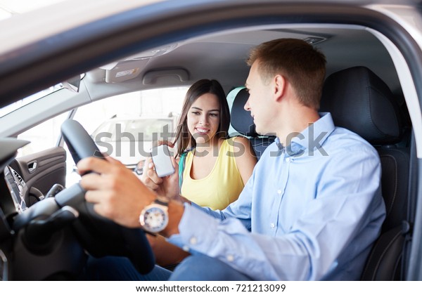 Buying a dream car. Portrait\
of a beautiful young couple buying a new car at the dealership\
handsome man laughing and hugging his wife while woman is showing\
thumbs up