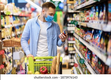 Buying Food. Portrait Of Young Man Wearing Medical Face Mask Shopping Groceries In Hypermarket, Standing With Trolley Cart Full Of Products In Shop Indoors, Holding Glass Jar Of Sauce, Checking Label