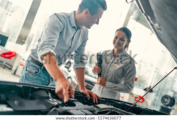 Buying my first car. Young car saleswoman\
standing at the dealership telling about the features of the car\
under the hood to the handsome\
man.