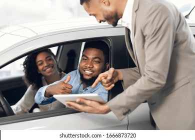 Buying New Car. Happy Black Couple Gesturing Thumbs Up Talking With Seller Approving New Automobile In Dealership Store