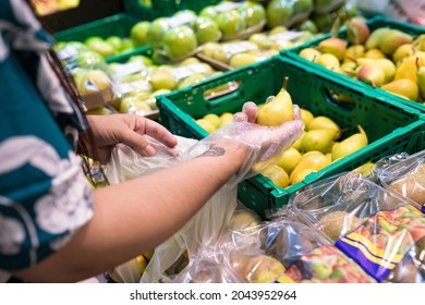 Buying Pears In A Supermarket. Woman Picking Up A Pear With Her Right Hand To Put It In A Bag. She Is Wearing Transparent Plastic Gloves For Hygienic Reasons. Healthy Eating Concept.