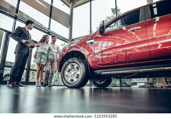 Buying their first car together. Bottom\
view of young car salesman standing at the dealership telling about\
the features of the car to the\
customers