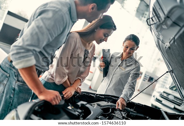 Buying their first car together. Young car\
saleswoman standing at the dealership telling about the features of\
the car under the hood to the\
customers.