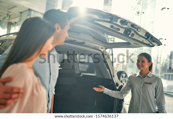 Buying their first car together. Young car\
saleswoman standing at the dealership telling about the features of\
the car to the\
customers.