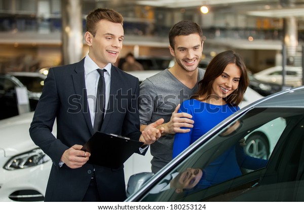 Buying their first car together. Handsome young\
car salesman standing at the dealership telling about the features\
of the car to the\
customers