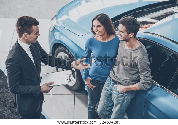 Buying their first car together. High angle\
view of young car salesman standing at the dealership telling about\
the features of the car to the\
customers