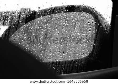 A B&W photo of a car's side mirror through the fogged up window, covered in raindrops, a dark mass in the lower corner of the photo. 