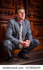 By The Door. Young Man Wearing Gray Suit, Neck Tie, Black Leather Shoes, Squatting By Office Door In New York City, Looking Up, Thinking. 
