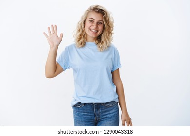 Bye see ya. Portrait of cute friendly and positive nice girl with blond hair and blue eyes smiling pleasant as waving palm in greeting or hello gesture being welcoming and nice over white wall