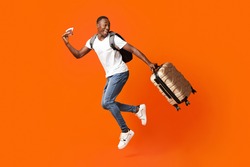 Bye-bye, Im Going Vacation. Young Happy Black Man With Suitcase, Backpack, Passport And Flight Tickets Jumping Up Orange Studio Background. Excited African American Guy Running And Looking Back