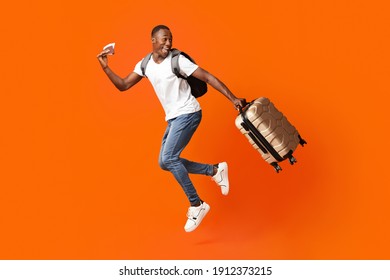 Bye-bye, Im going vacation. Young happy black man with suitcase, backpack, passport and flight tickets jumping up orange studio background. Excited african american guy running and looking back