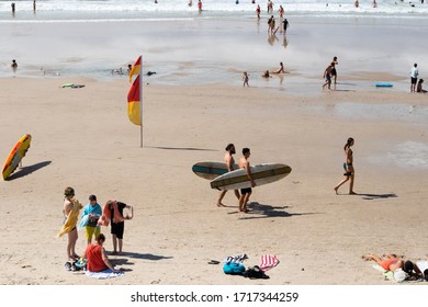 Byron Bay, Australia; March 2020: Beach Day: Two Young Men Carrying Surf Table Boards, Family Together, Kids Playing, People Walking, Surf Rescue Table On The Sand. Main Beach, Byron Bay, Australia