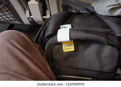 Cabin bag on a flight with tag for carry-on size guarantee fit between legs and under seat in front - Shutterstock ID 2169509993