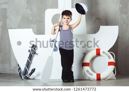 Cabin boy in striped t-shirt waving sailor cap in front of stylized ship, studio shot