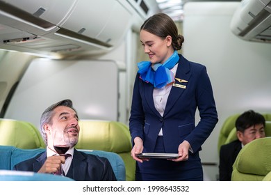 Cabin Crew Or Flight Attendant Talking With Businessman While Serving Red Wine To Him In Business Class Passenger Zone After Airplane Taking Off 