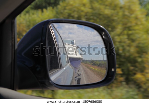 Cabin motor boat towing on car\
trailer, close up view in reflection in right side car rear mirrow\
on blue sky and green field background, travel with private\
boat