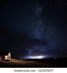 A Cabin With The Porch Light On And Fencing In The Fields With The Milky Way In The Starry Night Above Along With Lightning And Storm Clouds In The Distance Near Zion National Park In Utah.