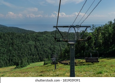 cable car construction with empty seats among green trees, grass field in forest on hill, sunny summer blue sky with white clounds, sea on horizon, sun light