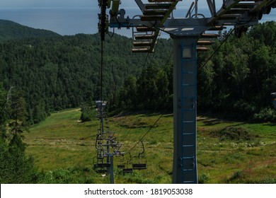 cable car construction with empty seats among green trees, grass field in forest on hill, sunny summer blue sky with white clounds, sea on horizon