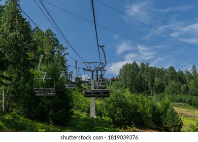 cable car construction with with wires and empty seats among green trees, grass field in forest on hill, summer sun light, blue sky with white clouds