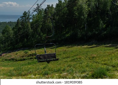 cable car empty seat among coniferous trees in green forest on hill in mountains, sunny summer blue sky with white clounds, baikal lake on horizon