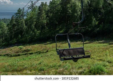 cable car empty seat among trees, grass field in green forest on hill, sunny summer blue sky with white clounds, baikal lake on horizon