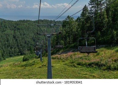 cable car empty seats among trees, grass field in green forest on hill, sunny summer blue sky with white clounds, baikal lake on horizon