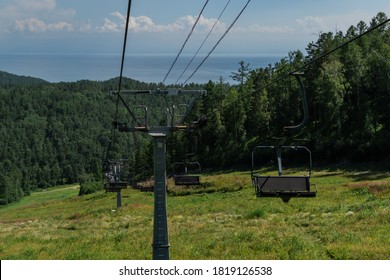 cable car empty seats among trees, grass field in green forest on hill, sunny summer blue sky with white clounds, sea on horizon