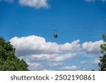 A cable car is flying through the sky above a forest. The sky is mostly clear, with a few clouds scattered throughout. The cable car is suspended high above the trees. The scene is peaceful