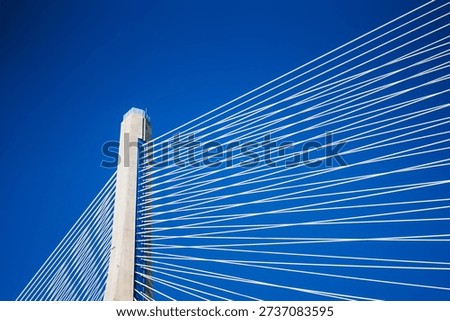 Cables creating geometric patterns connect to a concrete pylon against a bright blue sky