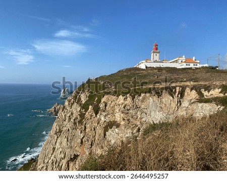 Cabo da Roca lighthouse perched on dramatic limestone cliffs. Portugal's westernmost point overlooks Atlantic Ocean. Red-topped beacon stands amid white buildings against azure sky.