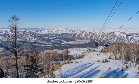 The cabs of the funicular move along the ropes over the snow-covered valley. Bare trees and firs are visible below. A picturesque mountain range in the distance. Altai. Manzherok Ski Resort - Shutterstock ID 2206092289