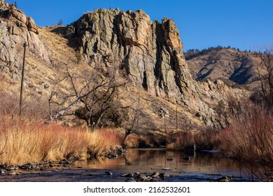 Cache la Poudre canyon as it approaches Fort Collins, CO.  Huge rock bluff, blue sky and trees along white clouds.  Rocky foreground.  Scenic area with blue sky, clear blue water and river rocks.