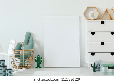 Cactus Shaped Pillow Placed In Wicker Basket Standing Next To Mockup Poster With Place For Your Photo In White Kid Room Interior With Cupboard And Rocking Horse