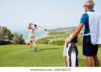 Caddy watching woman tee off on golf course overlooking ocean