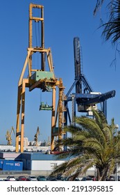 Cadiz, Spain. May 22nd 2022. Two Shipping Container Cranes Stand At A Dock With A Busy Car Park In The Foreground. Blue Sky And Tow Huge Cranes Dominate The Scene.