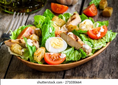 Caesar salad with croutons, quail eggs, cherry tomatoes and grilled chicken in wooden plate on dark rustic table