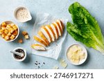 Caesar salad setup: fresh lettuce, grilled chicken, toasted croutons, grated Parmesan, peppered dressing, culinary delight. Blue background. Top view.