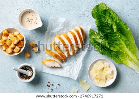 Caesar salad setup: fresh lettuce, grilled chicken, toasted croutons, grated Parmesan, peppered dressing, culinary delight. Blue background. Top view.