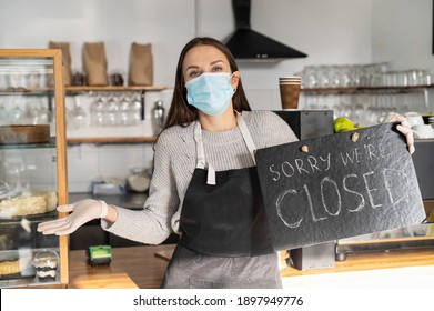 Cafe, Bar Closed Due Outbreak Lockdown, Female Wearing Medical Mask And Apron Looks At Camera, Holds A Closed Sign And Shrugs Shoulders. Small Business Crisis Concept