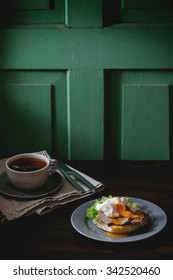 Cafe Breakfast With Homemade Sandwich With Baked Meat And Soft-boiled Egg And Cup Of Hot Pocket Tea Over Dark Wooden Table Near The Window With Green Wall At Background. Dark Rustic Style.