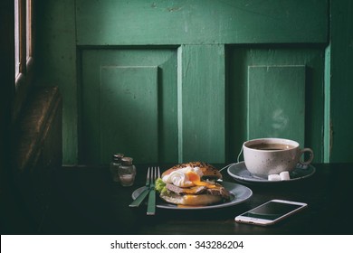 Cafe Breakfast With Homemade Sandwich With Baked Meat And Soft-boiled Egg And Cup Of Hot Pocket Tea Over Dark Wooden Table Near The Window With Turquoise Wall At Background. Dark Rustic Style.